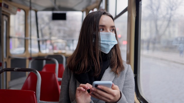 Porträt des jungen Mädchens in der schützenden medizinischen Gesichtsmaske, die Smartphone hält