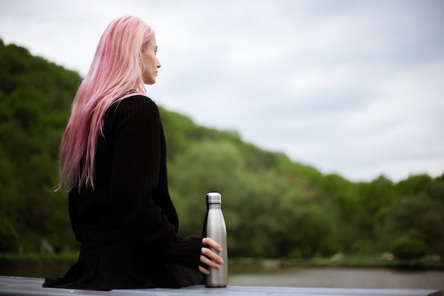Porträt des jungen Mädchens mit rosa Haaren, die im Park mit Thermoflasche in der Hand sitzen.