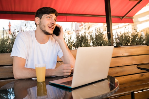 Porträt des jungen Mannes, der am Telefon spricht und laptp verwendet, während er in einem Café draußen sitzt. Kommunikationskonzept.