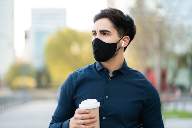 Porträt des jungen Mannes, der eine Tasse Kaffee hält, während draußen auf der Straße steht. Neues normales Lifestyle-Konzept.