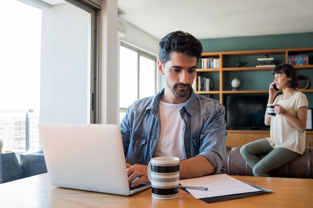 Porträt des jungen Mannes, der mit einem Laptop von zu Hause aus arbeitet, während Frau am Telefon spricht