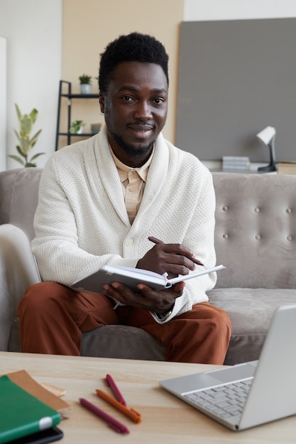 Porträt des jungen Mannes, der vorne lächelt, während er auf dem Sofa mit Büchern sitzt, die er zu Hause studiert