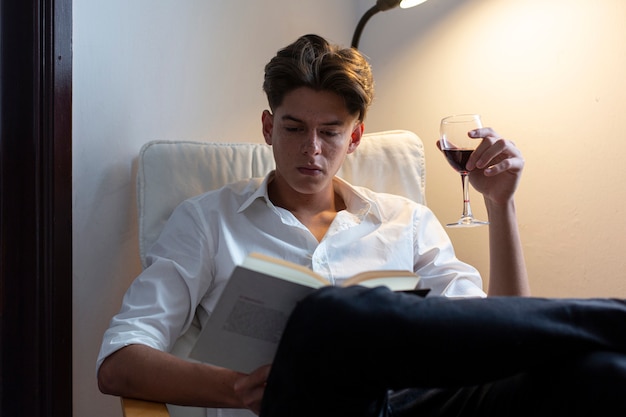 Porträt des jungen Mannes im weißen Hemd, das Wein trinkt und ein Buch zu Hause liest