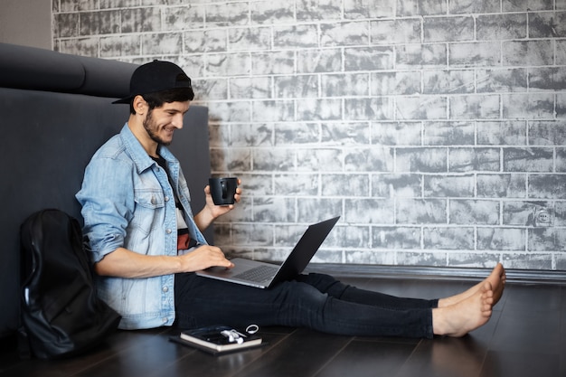 Porträt des jungen Mannes in der Jeansjacke und in der schwarzen Kappe, die auf dem Boden sitzen und am Laptop arbeiten, unter Verwendung des Smartphones, schwarze Tasse Kaffee.