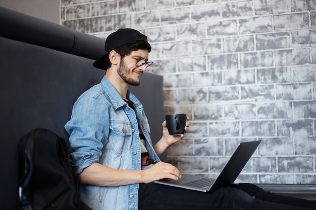 Porträt des jungen Mannes in der Jeansjacke und in der schwarzen Kappe, die auf dem Boden sitzen und am Laptop arbeiten, unter Verwendung des Smartphones, schwarze Tasse Kaffee.