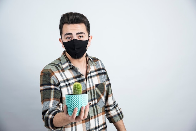 Porträt des jungen Mannes in der schwarzen Maske, die kleinen Kaktus im blauen Topf über weißer Wand hält.