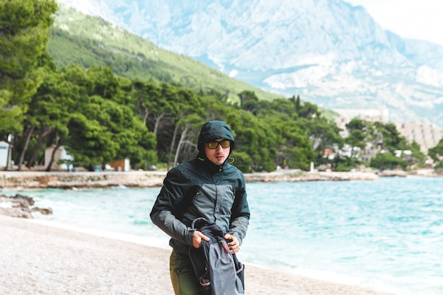 Porträt des jungen Mannes mit Regenmanteljacke am Strand im Sommer, der vom Sturm wegläuft, der vom Meer sich nähert. Vorhersage des Klimawandels für schlechtes Wetter und kalte Luft.