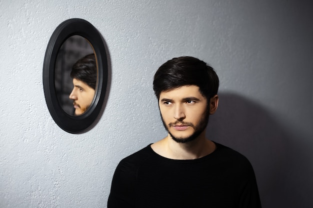 Porträt des jungen nachdenklichen Mannes nahe seinem Spiegelbild auf ovalem schwarzem Spiegel auf grauer strukturierter Wand.