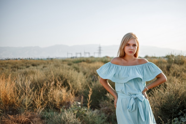 Porträt des jungen schönen kaukasischen blonden Mädchens im hellblauen Kleid, das auf dem Feld mit sonnengetrocknetem Gras während des Sonnenuntergangs steht