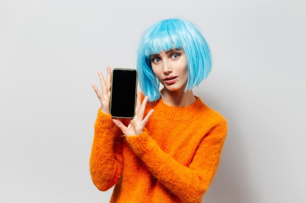 Porträt des jungen schönen Mädchens mit dem blauen Haar, das Smartphone in den Händen auf Hintergrund des hellgrauen hält; trägt einen orangefarbenen Pullover.