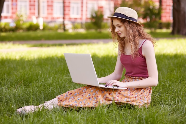 Porträt des jungen Teenagers, der auf grünem Gras im Park mit tragbarem Computer auf Beinen sitzt, Sommertag im Freien verbringend, Laptop und kabelloses Internet für das Schreiben der Kursarbeit verwendet. Studentenkonzept.