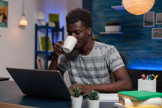 Porträt des jungen Unternehmers des Afroamerikaners, der am Schreibtisch sitzt und Marketingideen schreibt, die Finanzdiagramm auf Laptop-Computer analysieren. Mitarbeiter trinken Kaffee, während sie von zu Hause aus arbeiten