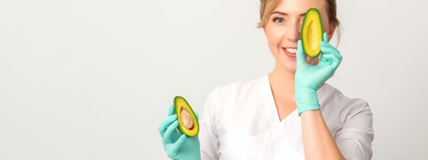 Porträt des jungen weiblichen Ernährungswissenschaftlerdoktors mit dem schönen Lächeln, das an der Kamera posiert, die das Auge hinter der halben Avocado auf weißem Hintergrund versteckt, Kopienraum. Vorteile der richtigen Ernährung.