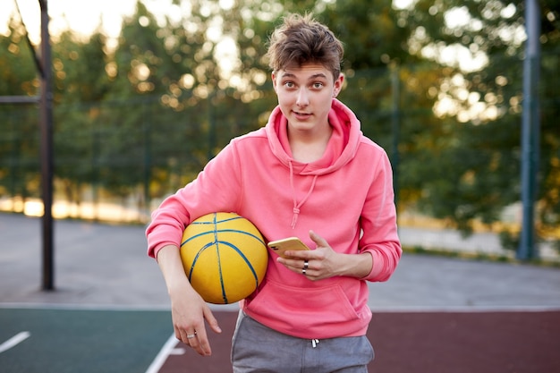 Porträt des kaukasischen Basketballerjungen mit Smartphone und Ball in den Händen
