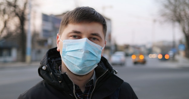 Porträt des kaukasischen verärgerten kranken Mannes in der medizinischen Maske, die an der Straße steht und schaut. Nahaufnahme des ungesunden Kerls, der Schutz vor Coronavirus in der Stadt trägt. Pandemie-Konzept. Infizierte Person.