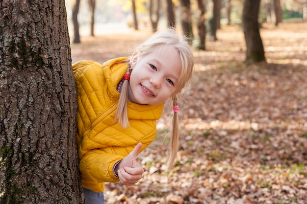 Porträt des Kindermädchens schaut hinter Baum heraus und zeigt Daumen hoch