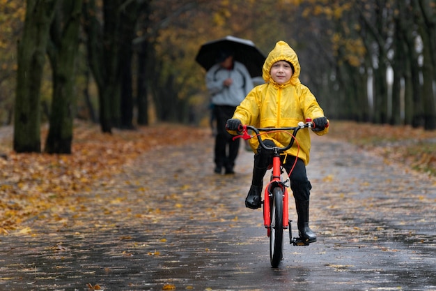 Porträt des Kindes auf dem Fahrrad im Regen. Junge im gelben Regenmantel reitet im Park