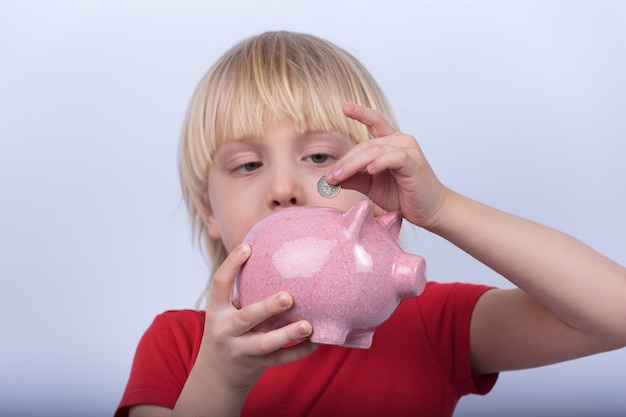 Porträt des Kindes auf weißem Hintergrund mit Sparschwein in der Hand. Junge steckt Münze in Sparschwein.