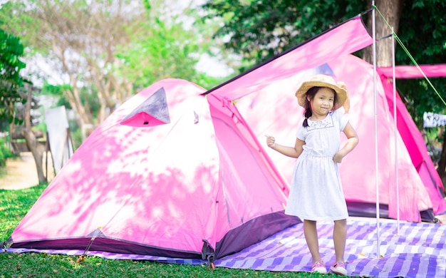 Porträt des kleinen asiatischen Mädchens, das Hut trägt, der mit Zelten steht, während Camping geht