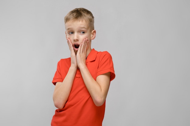 Porträt des kleinen blonden überraschten überraschten kaukasischen Jungen im orange T-Shirt mit offenem Mund auf grauem Hintergrund