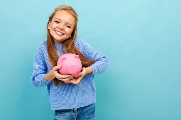 Porträt des kleinen kaukasischen Mädchens mit langen Haaren hält rosa Schwein Moneybox lokalisiert auf blauem Hintergrund