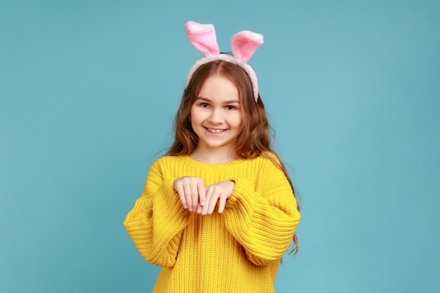 Porträt des kleinen Mädchens steht mit rosa Hasenohren, Kind hat Spaß beim Spielen, schaut in die Kamera und trägt einen gelben Pullover im Freizeitstil. Studioaufnahme im Innenbereich isoliert auf blauem Hintergrund.
