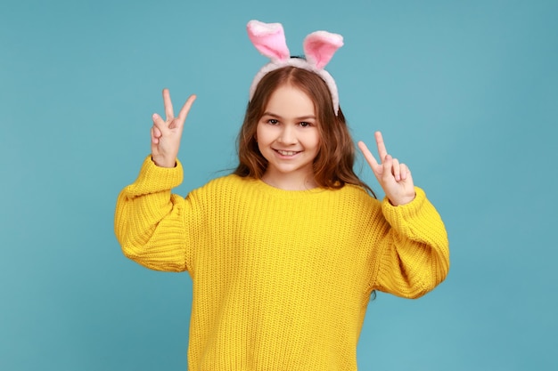 Porträt des kleinen Mädchens steht mit rosa Hasenohren, Kind hat Spaß, zeigt V-Zeichen zur Kamera und trägt gelben Pullover im lässigen Stil. Studioaufnahme im Innenbereich isoliert auf blauem Hintergrund.