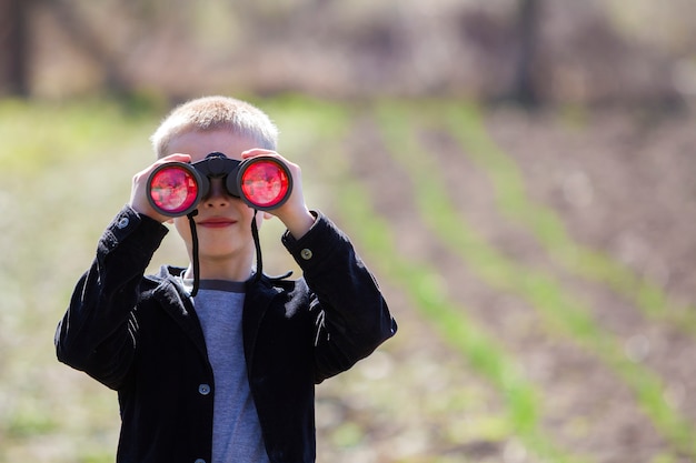 Porträt des kleinen netten hübschen netten blonden Jungen, der bedacht etwas durch Ferngläser im Abstand aufpasst