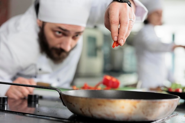 Porträt des Küchenchefs, der mit Gourmetgericht aus roter Paprika zum Abendessen in der professionellen Küche des Restaurants garniert. Gastronomieexperte wirft beim Kochen frisches Bio-Gemüse in die Pfanne.