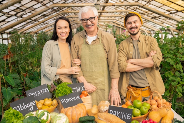 Porträt des lächelnden älteren Gärtners und seiner jungen multiethnischen Assistenten, die am Tisch mit reifem Gemüse am Bauernmarkt stehen