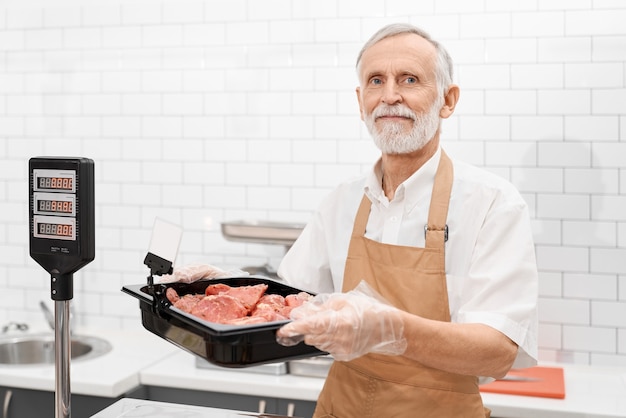 Porträt des lächelnden älteren männlichen Metzgers, der ein Stück frisches rotes rohes Fleisch in den Händen hält. Fröhlicher Mann hinter Zähler des Geschäfts, der Fleisch zeigt und Schüssel vom Kühlschrank auf Waage im Supermarkt setzt.