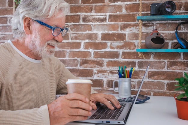 Porträt des lächelnden älteren Mannes, der zu Hause Laptop-Computer verwendet. Backsteinmauer im Hintergrund