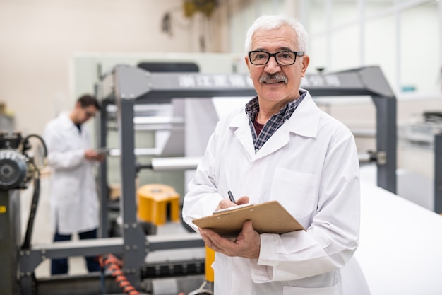 Porträt des lächelnden älteren Qualitätsinspektors im Laborkittel, der Notizen in der Zwischenablage bei Fabrik macht