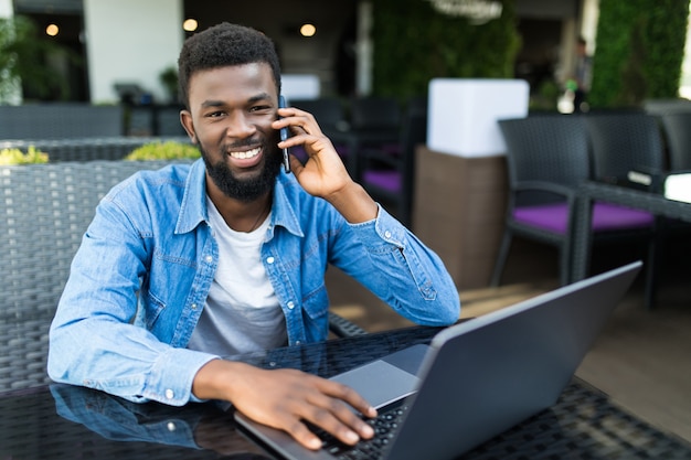 Porträt des lächelnden afrikanischen Mannes, der auf Handy spricht, während er an einem Café mit einem Laptop sitzt