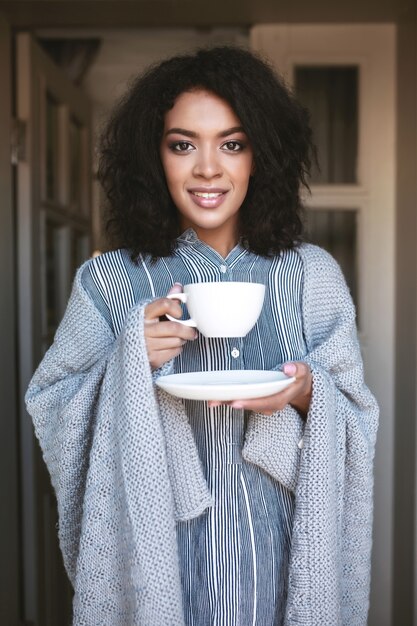 Porträt des lächelnden afroamerikanischen Mädchens eingewickelt im Plaid mit Tasse Kaffee in den Händen. Hübsche Dame mit dunklem lockigem Haar, das Kaffee trinkt