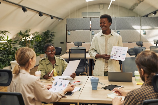 Porträt des lächelnden afroamerikanischen Mannes, der Datentabelle hält, während Bericht der Statistik und Leistung während des Geschäftstreffens im modernen Büro, Kopierraum gibt