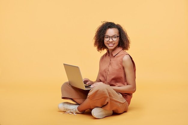Porträt des lächelnden afroamerikanischen Studentenmädchens mit dem gelockten Haar, das mit gekreuzten Beinen sitzt und Laptop gegen gelben Hintergrund verwendet