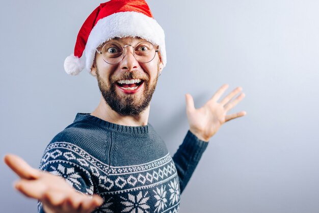 Porträt des lächelnden bärtigen Kerls, der Weihnachtsmütze, Brille und blauen Strickpullover mit leerer grauer Wand auf dem Hintergrund trägt