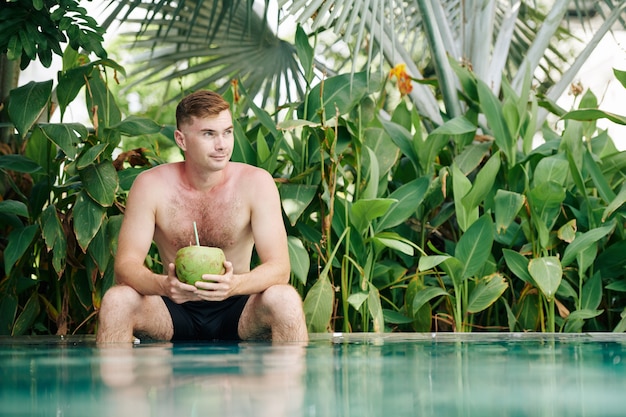 Porträt des lächelnden fot hemdlosen Mannes, der am Rand des Schwimmbades sitzt, Kokosnusscocktail trinkt und wegschaut