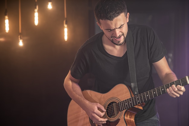 Porträt des lächelnden Gitarristen im schwarzen T-Shirt, das Akustikgitarre auf der dunklen Wand des unscharfen Studios spielt.