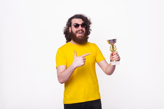 Porträt des lächelnden Hipster-Mannes mit dem lockigen Haar, das auf Championpokal zeigt