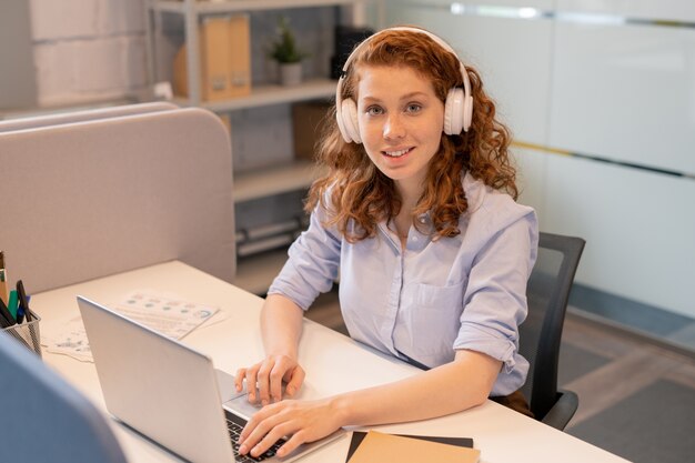 Porträt des lächelnden hübschen rothaarigen Dame-Managers in den drahtlosen Kopfhörern, die am Schreibtisch im Coworking Space sitzen und E-Mail auf Laptop beantworten