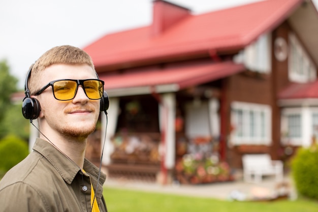 Porträt des lächelnden jungen bärtigen Kerls in der gelben Brille lässig gekleidet, die Musik online durch moderne Kopfhörer hört. Gemütliches Landhaus auf verschwommener Straße.