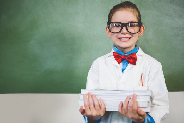 Porträt des lächelnden Mädchens, das mit einem Stapel Bücher im Klassenzimmer steht