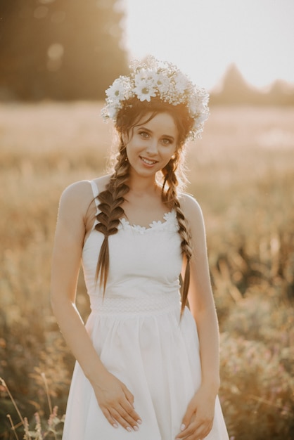 Porträt des lächelnden Mädchens im weißen Kleid mit Blumenkranz und Borten im Sommer bei Sonnenuntergang auf dem Gebiet