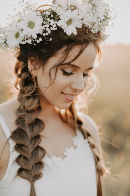 Porträt des lächelnden Mädchens im weißen Kleid mit Blumenkranz und Borten im Sommer bei Sonnenuntergang auf dem Gebiet