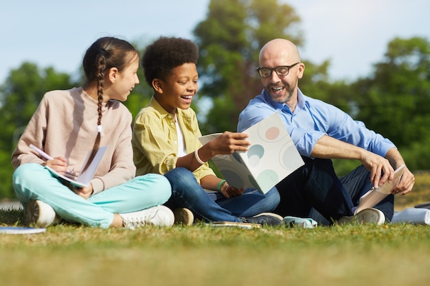 Porträt des lächelnden männlichen Lehrers in voller Länge, der mit Kindern spricht, während er auf grünem Gras sitzt und Klasse im Freien im Sonnenlicht genießt