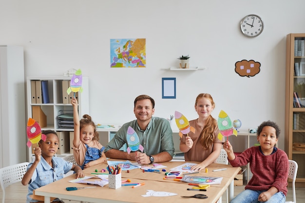 Porträt des lächelnden männlichen Lehrers mit multiethnischer Gruppe von Kindern, die Bilder von Weltraumraketen zeigen, während Kunst- und Handwerksunterricht in der Vorschule oder im Entwicklungszentrum genießen