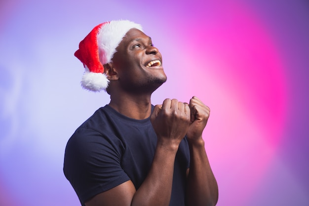 Porträt des lächelnden Mannes des positiven Afroamerikaners in Sankt-Hut und beiläufigem T-Shirt auf buntem Hintergrund, Weihnachtszeit. Winterferien und emotionales Menschenkonzept.