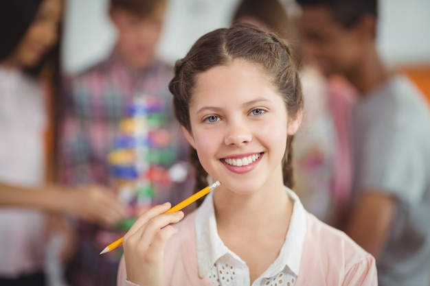 Porträt des lächelnden Schulmädchens, das mit Bleistift im Klassenzimmer steht